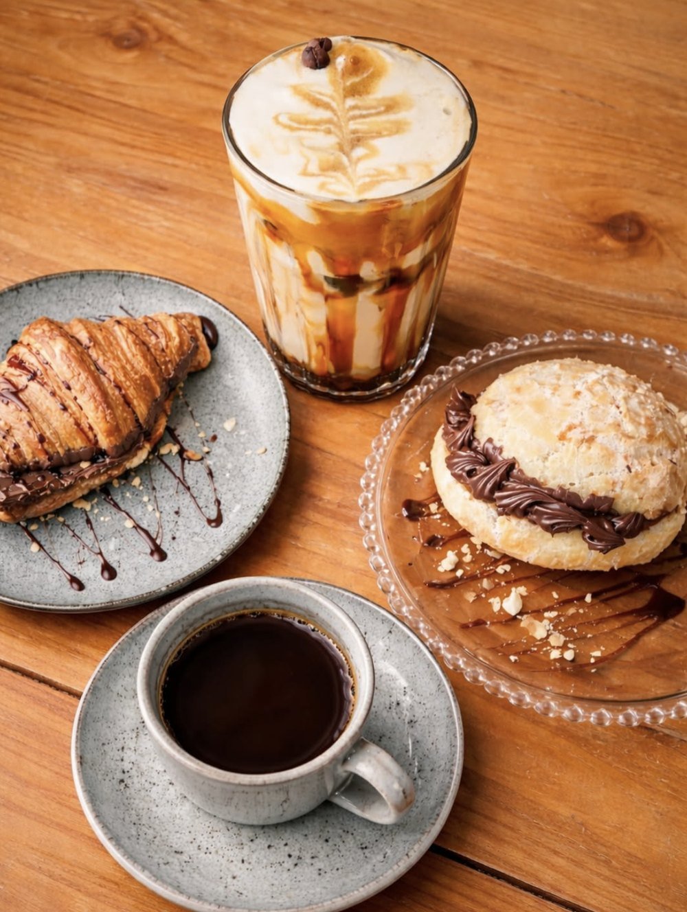 Café gelado com caramelo, croissant com Nutella, pão de queijo recheado e café coado sobre mesa de madeira